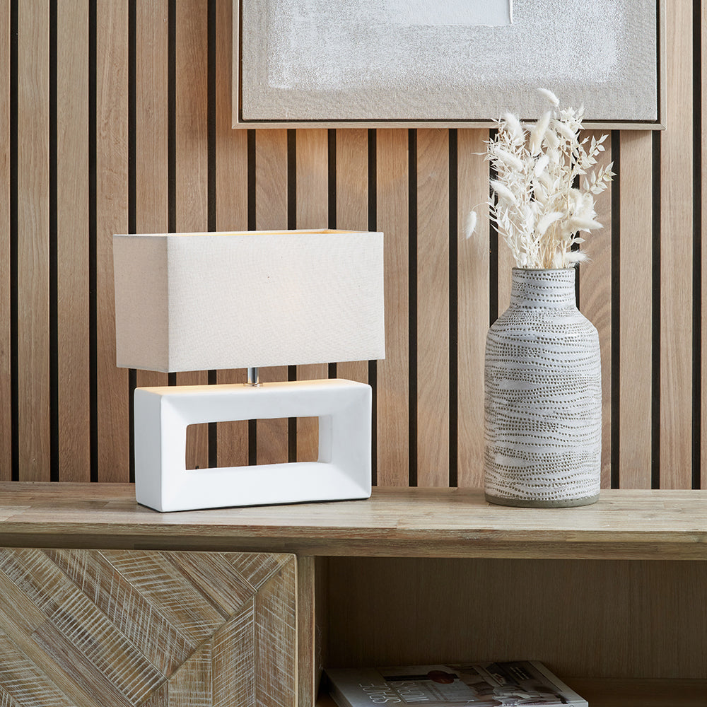 A modern white ceramic table lamp with a rectangular base and a neutral linen shade, placed on a wooden surface, with a decorative vase next to it.