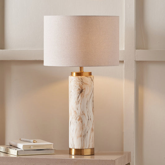 A ceramic table lamp with a natural stone effect finish and a gold-colored base, paired with a beige linen shade, placed on a side table in a home setting. 