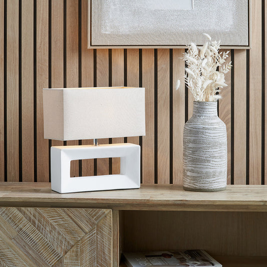 A modern white ceramic table lamp with a rectangular base and a neutral linen shade, placed on a wooden surface, with a decorative vase next to it. 