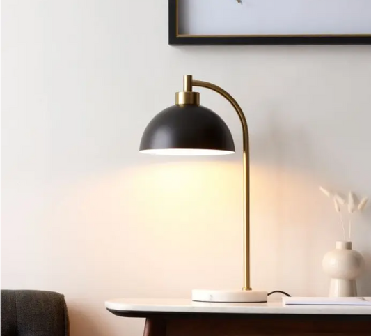 A modern matte black table lamp with a brass arched steel frame and a white marble base, turned on, providing warm light, placed on a desk. 