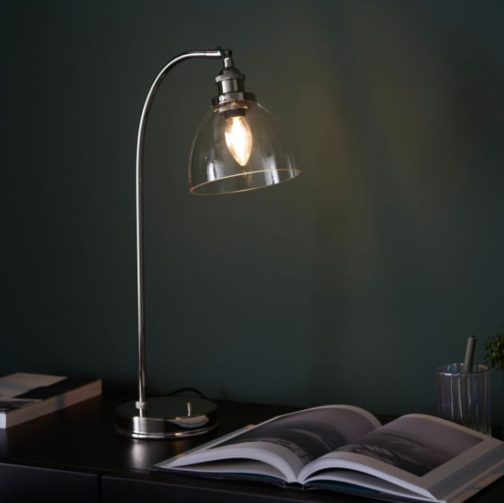 A modern bright nickel & clear glass table lamp with an arched design and a dome shade, turned on, providing a warm glow, placed on a table next to an open book -.