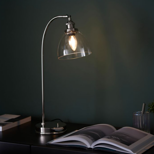 A modern bright nickel & clear glass table lamp with an arched design and a dome shade, turned on, providing a warm glow, placed on a table next to an open book -.