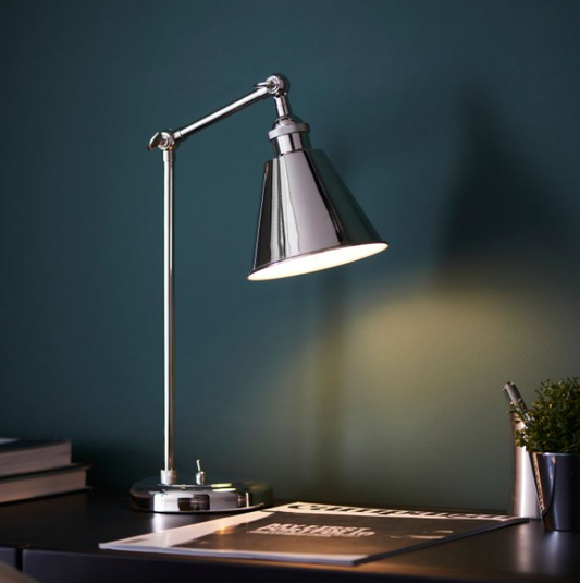 A bright nickel desk lamp with an adjustable arm and a conical shade, placed on a desk next to some books and a pen. 