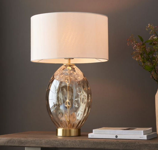 An elegant champagne glass table lamp with a textured base and an off-white fabric shade, placed on a wooden surface. 