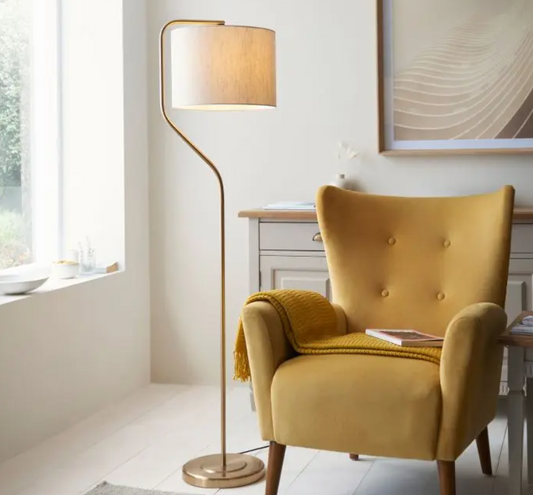 An elegant brushed aged brass floor lamp with a fabric shade, placed next to a yellow armchair in a room with a wooden floor and a picture on the wall.